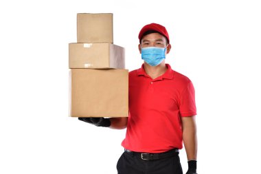 Kırmızı üniformalı, tıbbi maskeli, koruyucu eldivenli mutlu Asyalı teslimatçı beyaz arka planda karton kutu taşıyor. Teslimatçı çocuk paket teslimatı yapıyor. Güvenli teslimat.       