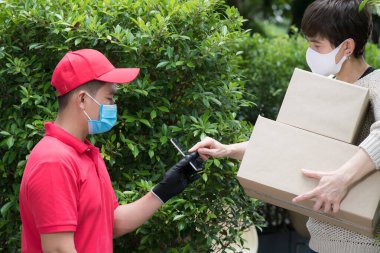 Yüz maskesi takan Asyalı teslimatçı ve kırmızı üniformalı eldiven COVID-19 salgını sırasında alıcıya paket kutusu teslim ediyor.