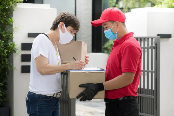 Yüz maskesi takan Asyalı teslimatçı ve kırmızı üniformalı eldiven COVID-19 salgını sırasında alıcıya paket kutusu teslim ediyor.