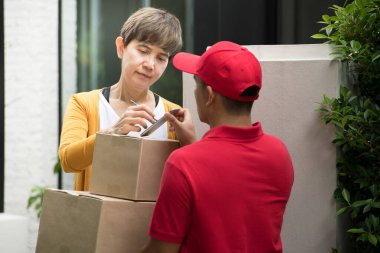 Kırmızı üniformalı Asyalı teslimatçı, paketin akıllı aygıtla alınması için alıcı imzasıyla paket kutusunu evdeki kadına teslim ediyor.