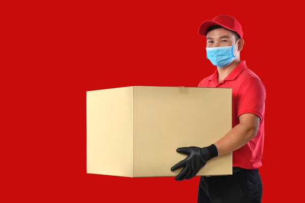 身穿红色制服 戴着医疗面罩 戴着防护手套的快乐的年轻亚洲送货员手拿着红底纸盒 送货的人把包裹送来 Covid 19爆发期间 — 图库照片