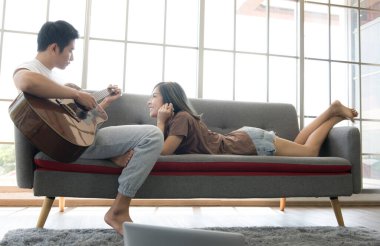 Genç Asyalı çift kanepede oturur ve evlerinde gitar çalarlar.