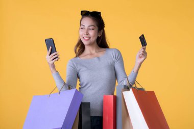 Akıllı telefonu ve kredi kartıyla sarı arka planda izole edilmiş renkli alışveriş çantaları olan güzel Asyalı genç bir kadın.