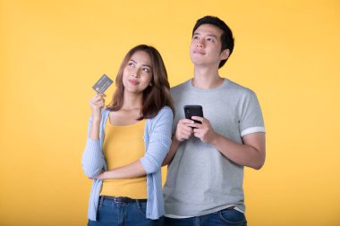 Genç Asyalı çift kredi kartı ve akıllı telefon tutarken sarı arka planda izole edilmiş bir yere bakıyor.