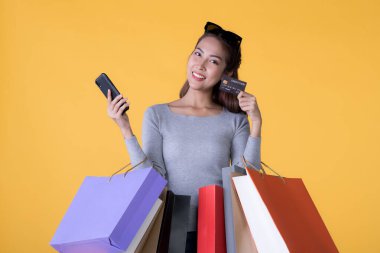 Akıllı telefonu ve kredi kartıyla sarı arka planda izole edilmiş renkli alışveriş çantaları olan güzel Asyalı genç bir kadın.