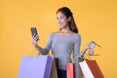 Renkli alışveriş çantaları ve akıllı telefonları olan güzel Asyalı genç bir kadın sarı arka grupta izole edilmiş.