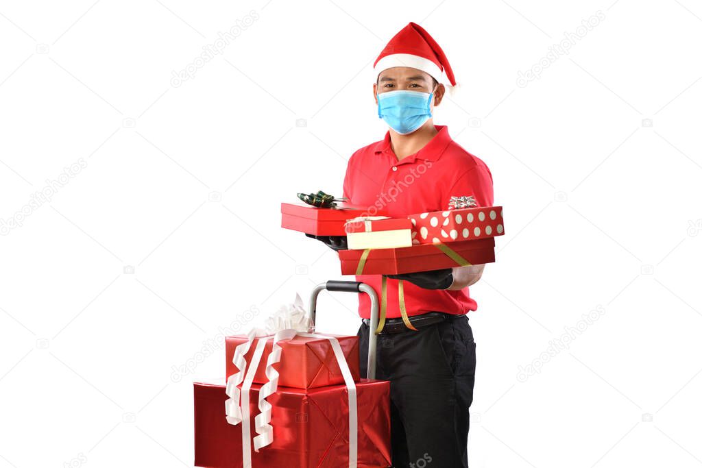 Happy young Asian delivery man in red uniform, medical face mask, protective gloves, Christmas hat carry boxes of presents in hands isolated on white background during the COVID-19 outbreak and Christmas festivities