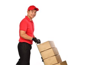 Kırmızı üniformalı genç Asyalı teslimatçı beyaz arka planda kutuları olan bir el arabasını itiyor. Teslimatçı bir paket teslimatı yapıyor..