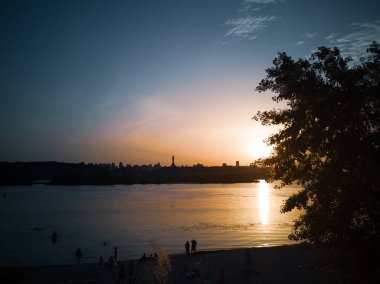 İnsanlar bir yaz akşamı Kyiv 'deki Dnipro nehrinin sol kıyısında yüzüyor ve plajda dinleniyorlar. Anavatanı olan şehir manzarası güneşin battığı yerde, sağ kıyıda..