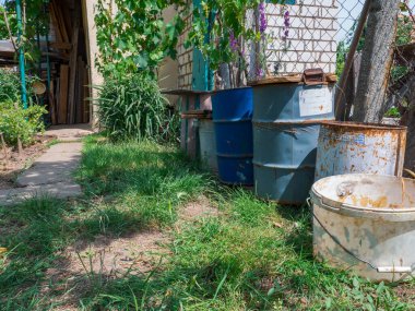 Köy evinin arka bahçesinde ahşap kapakları ve plastik kovaları olan paslı metal variller..
