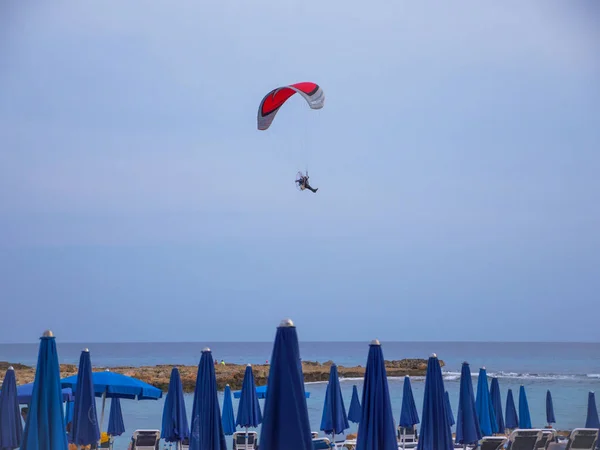 Powered paraglider wtih red wing flying high in the evening blue sky above Nissi beach with blue umbrellas in Ayia Napa, Cyprus. — Stock Image Powered Paraglider Wtih Red Wing Flying High Evening Blue Sky Stock Picture