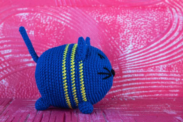 Soft knitted toy cat. Round. Of blue color. In the technique of amigurumi. On a red textural background. Looks to the right.