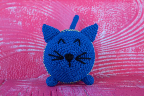 Soft knitted toy cat. Round. Of blue color. In the technique of amigurumi. On a red textural background. Looks straight.