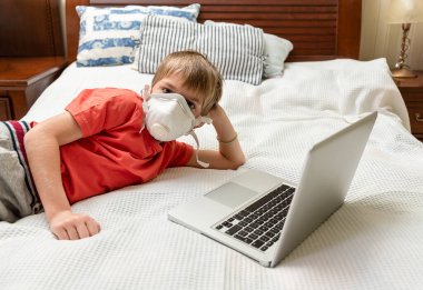 Tıbbi maskeli çocuk dizüstü bilgisayar kullanıyor ve kameraya bakıyor.. 