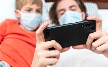Akıllı telefon tutan, tıbbi maske takan ve karantina sırasında evde oynayan iki kardeş..