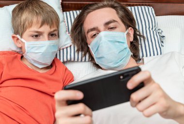 Tıbbi maske takan ve telefonla oynayan iki kardeş, Covid-19 karantinası sırasında evde izole edilmiş.. 