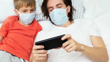 Tıbbi maske takan ve telefonla oynayan iki kardeş, Covid-19 karantinası sırasında evde izole edilmiş..