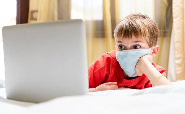 Evde dizüstü bilgisayarı olan tıbbi maskeli bir çocuk var. Covid-19 karantina, online okul.