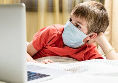 Evde dizüstü bilgisayarı olan tıbbi maskeli bir çocuk var. Covid-19 karantina, online okul.