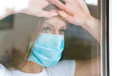Karantina sırasında pencereden dışarı bakan tıbbi maskeli kadın, Covid-19 salgını..