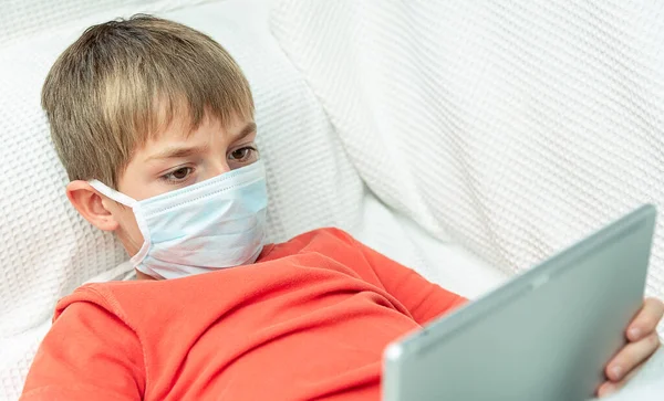 Tıbbi maskeli çocuk tablette oynuyor.
