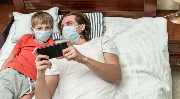 Tıbbi maske takan ve telefonla oynayan iki kardeş, Covid-19 karantinası sırasında evde izole edilmiş..