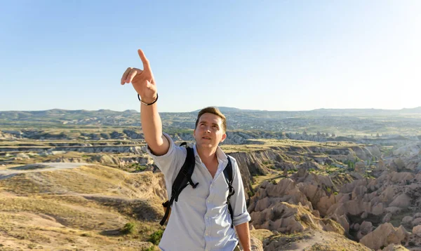 Young man with backpack pointing with right hand and standing against beautiful valley at Cappadocia. Bleisure concept