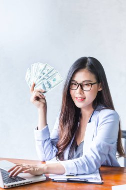 Güzel bir iş kadını ofis masasında dizüstü bilgisayar kullanarak oturuyor ve elinde banknotla çalışırken gülümsüyor ve bilgisayar kullanarak neşeli bir model oluyor. Kapalı