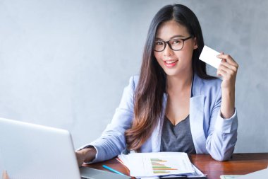 Güzel bir iş kadını ofis masasında dizüstü bilgisayar kullanarak oturuyor ve elinde kredi kartıyla çalışırken gülümsüyor ve bilgisayar kullanarak neşeli bir model oluyor. Kapalı