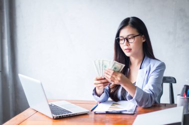 Güzel bir iş kadını ofis masasında dizüstü bilgisayar kullanarak oturuyor ve elinde banknotla çalışırken gülümsüyor ve bilgisayar kullanarak neşeli bir model oluyor. Kapalı