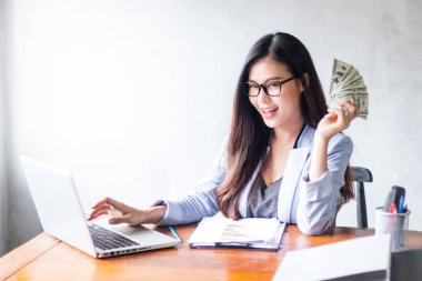 Güzel bir iş kadını ofis masasında dizüstü bilgisayar kullanarak oturuyor ve elinde banknotla çalışırken gülümsüyor ve bilgisayar kullanarak neşeli bir model oluyor. Kapalı