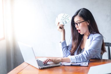 Güzel bir iş kadını ofis masasında dizüstü bilgisayar kullanarak oturuyor ve elinde banknotla çalışırken gülümsüyor ve bilgisayar kullanarak neşeli bir model oluyor. Kapalı