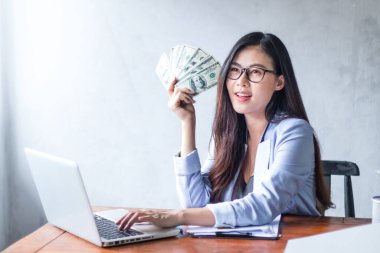 Güzel bir iş kadını ofis masasında dizüstü bilgisayar kullanarak oturuyor ve elinde banknotla çalışırken gülümsüyor ve bilgisayar kullanarak neşeli bir model oluyor. Kapalı