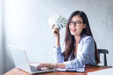 Güzel bir iş kadını ofis masasında dizüstü bilgisayar kullanarak oturuyor ve elinde banknotla çalışırken gülümsüyor ve bilgisayar kullanarak neşeli bir model oluyor. Kapalı