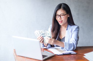 Güzel bir iş kadını ofis masasında dizüstü bilgisayar kullanarak oturuyor ve elinde banknotla çalışırken gülümsüyor ve bilgisayar kullanarak neşeli bir model oluyor. Kapalı