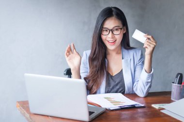 Güzel bir iş kadını ofis masasında dizüstü bilgisayar kullanarak oturuyor ve elinde kredi kartıyla çalışırken gülümsüyor ve bilgisayar kullanarak neşeli bir model oluyor. Kapalı