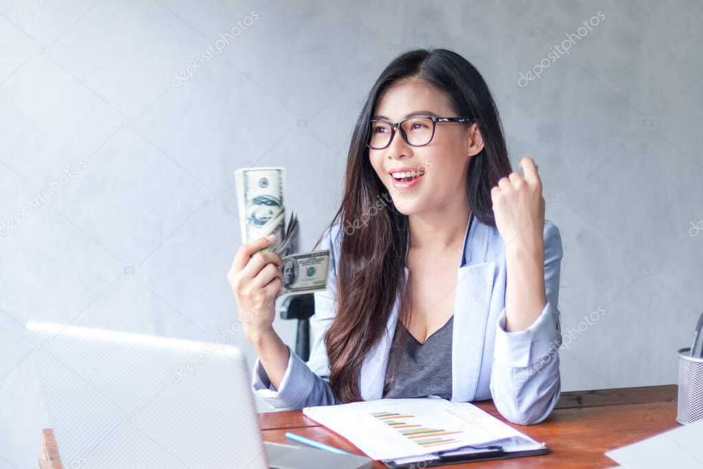 Beautiful business woman sitting at home office desk  using a laptop and smiling while working with banknote hold in hand, and and be attractive cheerful model using computer. Indoors