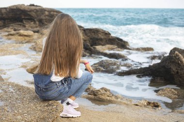 Genç güzel kıvırcık kız kot giymiş denize yakın kaya üzerinde oturuyor.