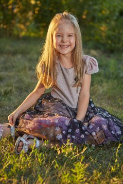 Küçük sevimli kız yaz bahçesinde çimlerin üzerinde oturuyor, gün batımında.