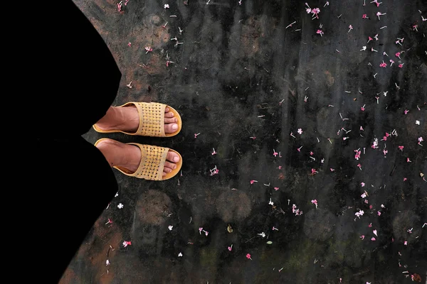 High view scene with feet of woman wear black pants and yellow plastic sandals stand in floor with tiny flower petal fall in garden