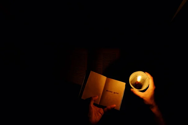 Miss you letter on notebook paper, woman hand hold candle with warm light, message for love far away on black background, simple concept in dark