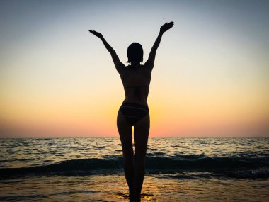 Denizde gün batımına karşı ince güzel bir kadının silueti. Batan güneşin ışınları yla sahilde duran bir kadının seksi çekici figürü. Sahildekız ellerini uzalar.  