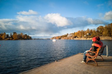 Sonbahar deniz manzarası. Baltık Denizi Körfezi. Iskandinavya. Hygge, ne oldu? Rahatla. Çift iskelede bir bankta oturur ve denize bakar. Gökyüzü, beyaz bulutlar, Kayalıklar, Ormanlık Adalar, çam ağaçları, ufuk. 