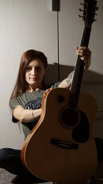 Dövmeli yeşil gözlü bir esmer elinde bir gitar tutuyor..