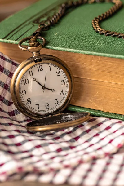 Pocket watch sitting on top of a plaid cloth with a book on the side. The time on the pocket watch is 3:49.