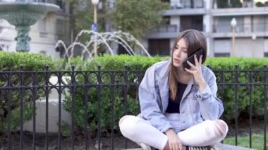 Genç kız arka planda çeşme ile bir bankta açık havada telefonda konuşurken