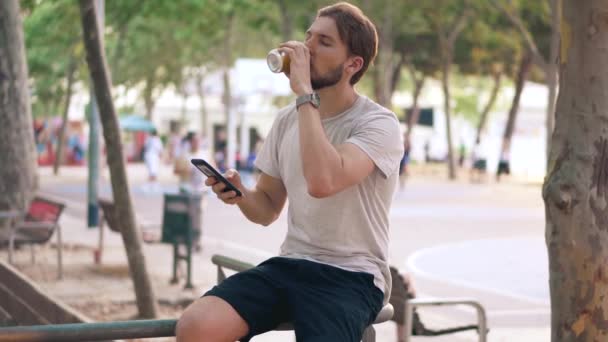 Молодой человек пьет пиво и проверяет смартфон в парке, сидя на рельсах — стоковое видео