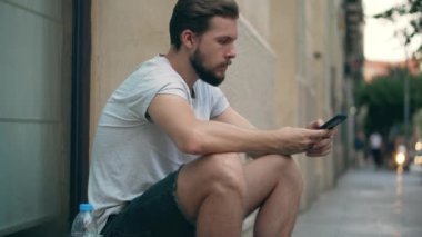 Sokakta oturan genç adam akıllı telefon kullanıyor.