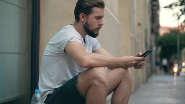 Молодой человек сидит на улице со смартфоном — стоковое видео