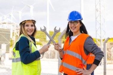 İki genç teknik mühendisin mutlu portresi kameraya bakıp gülümserken kaslarını metal İngiliz anahtarıyla elektrik ve elektrik santralinin rüzgar türbinlerine karşı esnetiyor.